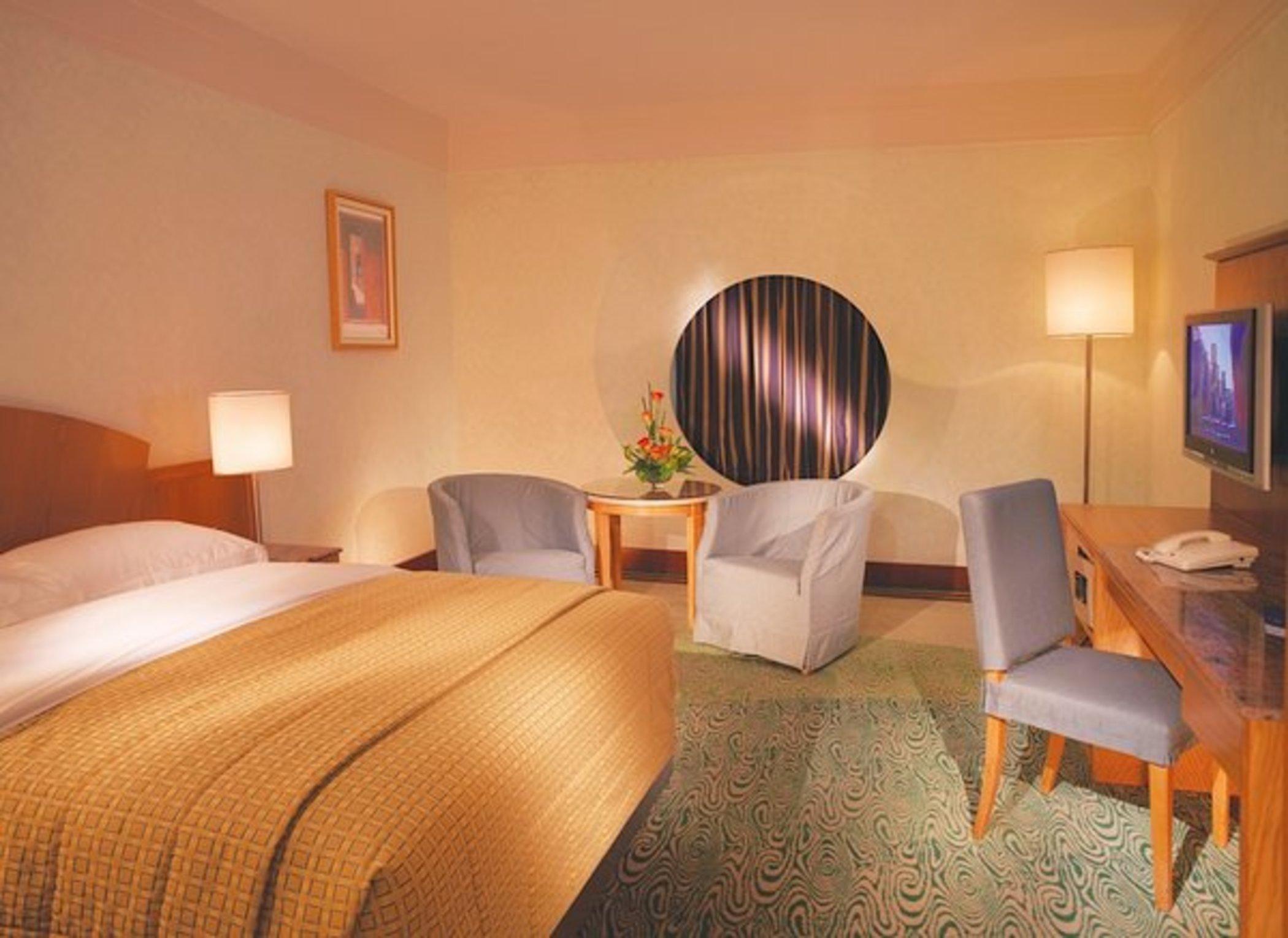 Dubai International Terminal Hotel Room photo The photo shows a cozy hotel room featuring a large bed with a patterned yellow bedspread. There are two upholstered chairs and a small table beside them. A round window is set into the wall, partially covered by curtains. A television is mounted on 