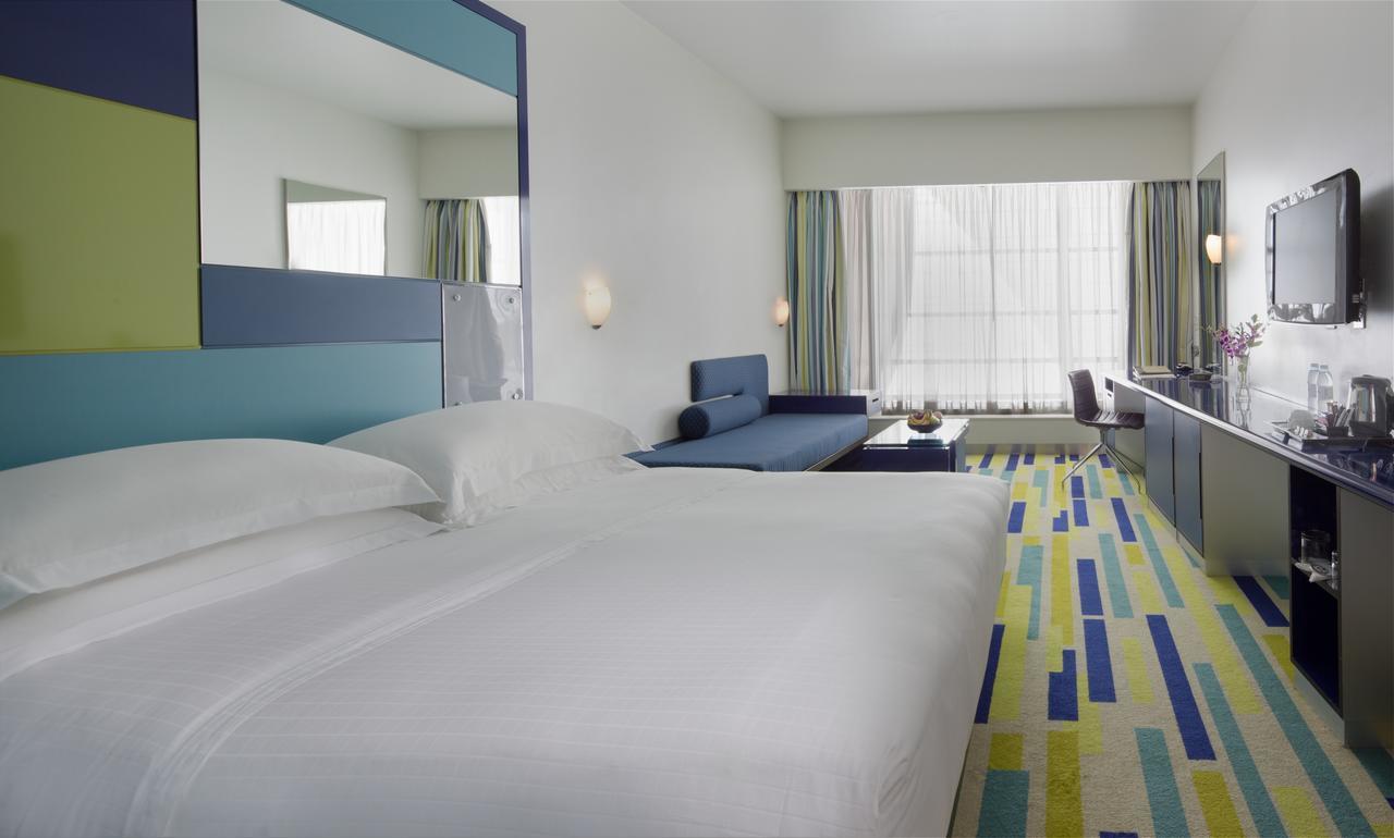 Dubai International Terminal Hotel Exterior photo The photo shows a modern hotel room featuring two large beds with white bedding. There is a colorful carpet with a geometric pattern in blue, green, and yellow. A large window lets in natural light, and light-colored curtains frame it. Along one wall