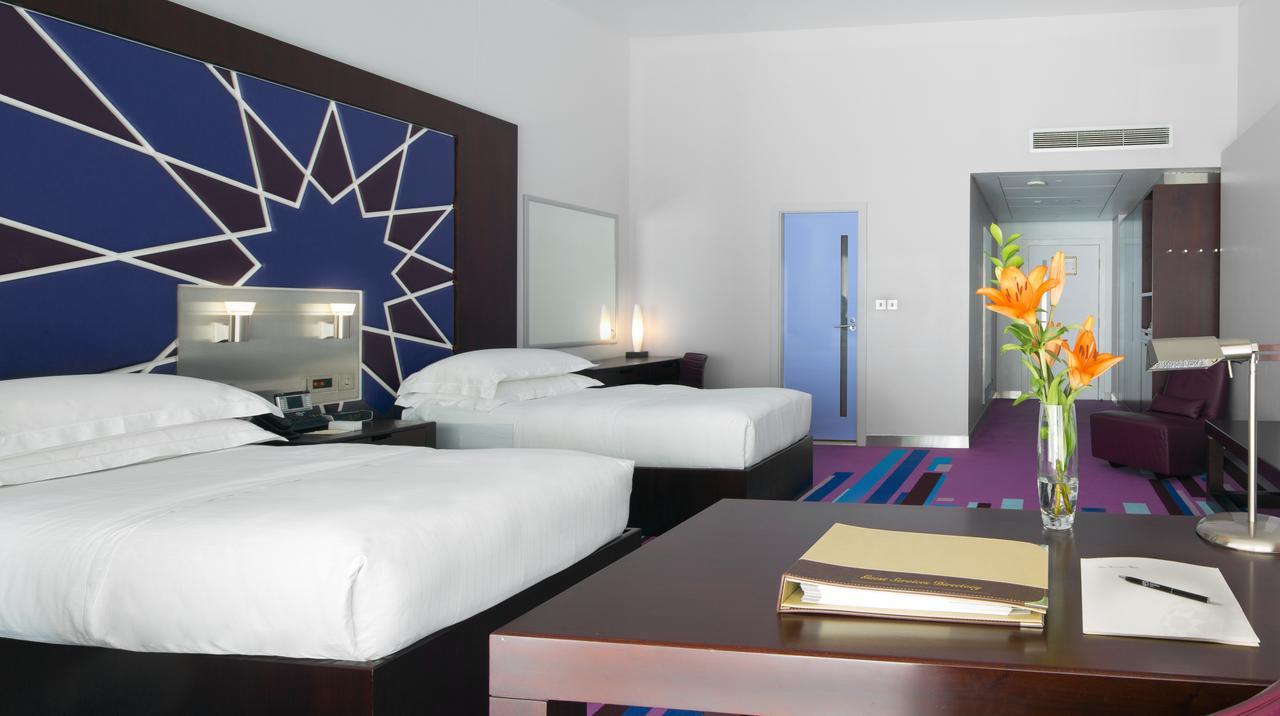 Dubai International Terminal Hotel Exterior photo The image shows a modern hotel room with two neatly made beds, white bedding, and dark wooden furniture. The walls have a striking geometric design, featuring a blue and white star shape. A small coffee table is in the foreground with a few items on 