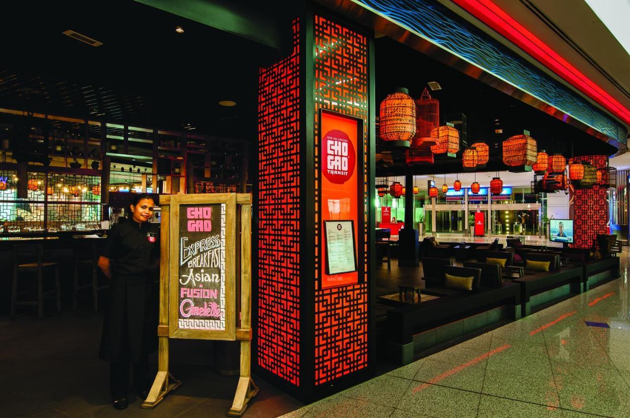 Dubai International Terminal Hotel Exterior photo The photo shows the entrance of a restaurant named "Gho Gho," featuring a prominent sign with the restaurant's name. A staff member in a black uniform stands beside a wooden sign that displays the restaurant's specialty, which is Asian fusion cuisine